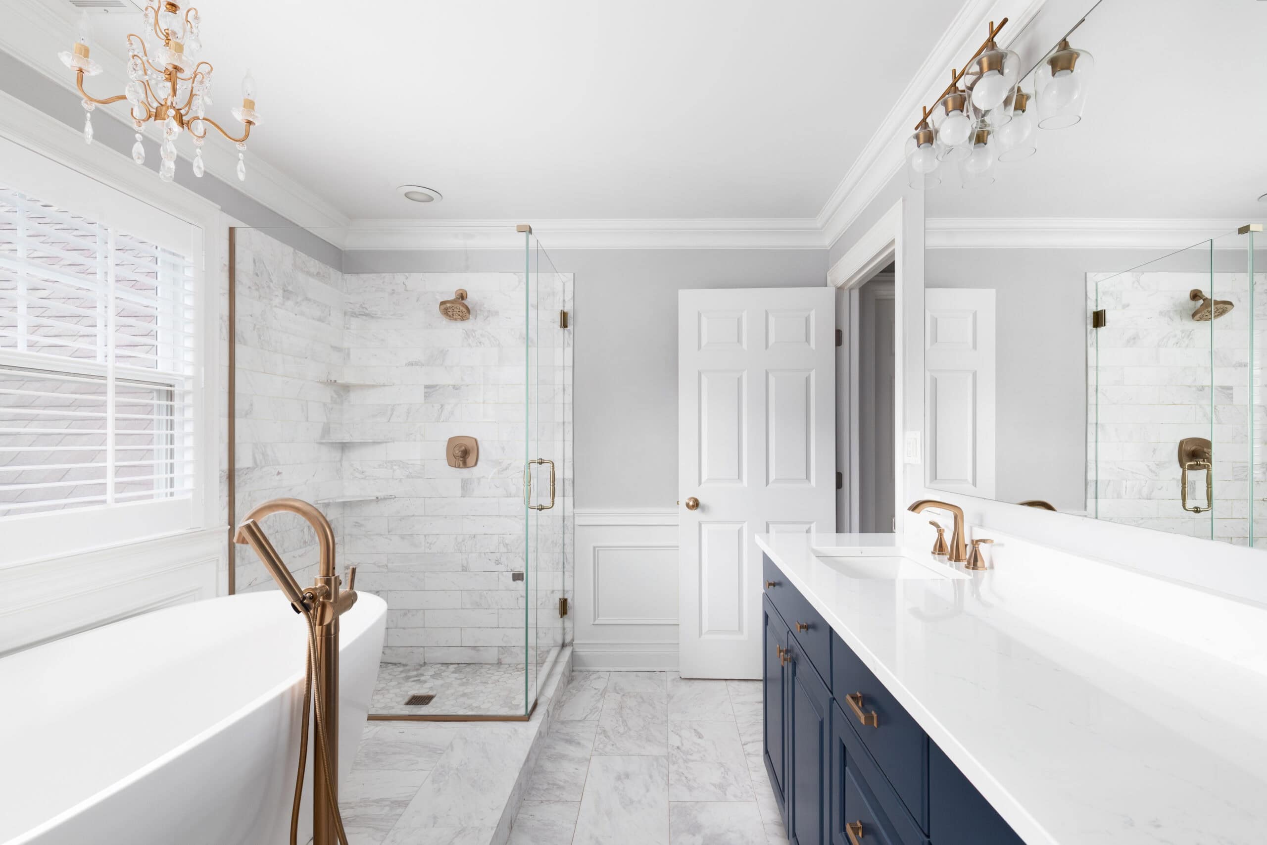 Modern bathroom with white marble walls, glass shower, and navy vanity with gold fixtures.